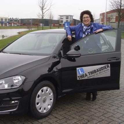 Thea Thibaudier bij haar lesauto met het bord Thea Thibaudier op het portier.